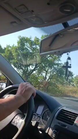 The video begins with a close-up of a driver's hand on the steering wheel of a car. The driver is wearing a watch on their left wrist. Through the windshield, a white police vehicle with a green stripe is visible, appearing to be stopped or moving slowly. The camera then pans to the right, revealing the interior of the car, including a rearview mirror and a black Nike baseball cap resting on the dashboard. The view outside the windshield shows a sunny day with a clear blue sky and trees lining the road. As the car moves, other vehicles, including cars and a van, are seen on the road ahead. The scenery consists of dry vegetation and trees, with some signs of infrastructure like utility poles and what appears to be a bridge in the distance. The video continues to show the car traveling on a paved road, with the landscape remaining consistent with a rural or semi-rural environment.
