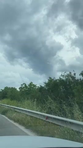 The video, filmed from inside a moving vehicle, shows a roadside scene under a heavily overcast sky. A white guardrail runs along the left side of the road, bordered by green grass and lush trees. The sky is filled with dark grey, voluminous clouds, with one particularly prominent, almost funnel-shaped cloud visible in the distance during the first few seconds. As the vehicle continues, the view progresses past the dense foliage. Towards the end of the clip, a red and white "No entry" sign (a red circle with a white horizontal bar) becomes briefly visible on the left side of the road, set back slightly from the guardrail. The location context provided is Aleko Konstantinovo, Bulgaria.