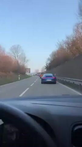 The view is from the driver's seat of a car, looking out at a blue Porsche ahead, traveling on a two-lane road with a line of other cars. There's some bare tree branches on the sides of the road, with some green foliage interspersed. A roadside billboard is visible to the left. There is a metal guard rail to the right of the road. The sky is clear and blue. The car is moving slowly in traffic.