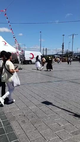 In the foreground, a paved square with a pattern of grey and darker grey stone tiles, the camera pans to the right. Two people, a young man and a young woman, walk across the square from left to right. The man wears a dark t-shirt with a pattern and dark pants, carrying a black backpack. The woman wears a light yellow t-shirt, white pants, and white sneakers, carrying a white plastic bag. In the middle ground, a white tent with a red crescent logo is set up. A white mobile unit, also with the red crescent logo and Turkish flags, is parked next to the tent. Red and white triangular flags are strung across the scene, and several people are visible in the background, some near the tent and others further away. The sky is blue with a few scattered white clouds.