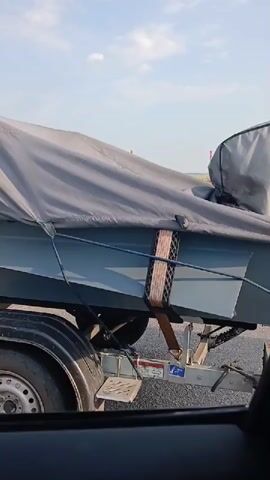 A gray boat is covered by a gray tarp and secured to a trailer with straps. The trailer is attached to a pickup truck. The scene appears to be outdoors on a paved surface, possibly a road or parking lot, under a partly cloudy sky. The camera perspective is from inside another vehicle.