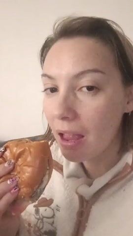 A woman is eating a hamburger in what appears to be an interior setting. She is fair-skinned with light brown hair and brown eyes. The woman is wearing a white, fuzzy, zip-up hoodie with a Snoopy design on the front. She is holding a hamburger with both hands and takes a large bite. The burger has a glossy bun and appears to contain meat and other fillings. Her fingernails are decorated with a colorful and patterned design. The background is a plain white wall.