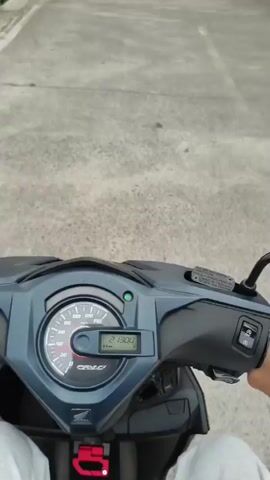 The video provides a first-person perspective of someone riding a scooter on a concrete road in Gadog, Indonesia. The initial frame shows the scooter's dashboard, with the odometer displaying "21300" and a green indicator light on. As the scooter begins to move, the scene reveals a narrow road lined with buildings, power poles, and overhead power lines under an overcast sky. On the left side of the road, a building features a sign that reads "WISMA PANGINAPAN HOEDILUHUR". Further along on the right, a green gate is visible with a "RUMAH INI DIJUAL" (This House For Sale) sign, surrounded by green foliage. The ride continues past more structures, including buildings with closed shutters, and a utility pole heavily laden with wires.