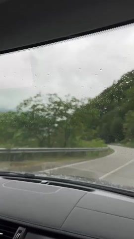 The video shows a car's dashboard and the view through the rain-streaked windshield. The vehicle is driving along a wet, winding road. Lush green trees and mountainsides flank the road, and the sky is overcast and gray. A metal guardrail is visible on the driver's side.