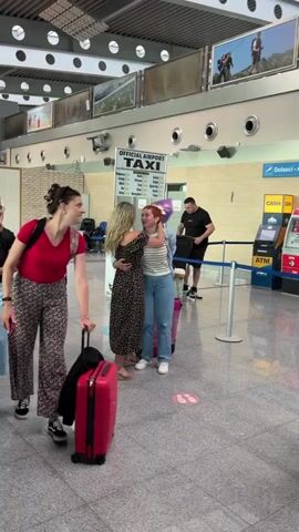 A group of people are gathered in what appears to be an airport terminal. In the foreground, a woman in a red shirt and floral pants is pulling a red suitcase with a black garment draped over it. To her left, another woman in a black shirt and blue pants is holding a phone and pulling a purple suitcase. Behind them, two women are embracing, one in a patterned dress and the other in a striped shirt and blue jeans. Both are holding luggage. Further in the background, several other individuals are visible, including a man in a black t-shirt standing near a taxi sign and a row of ATMs. The environment is bright and features architectural elements like a tiled floor, white ceiling with circular lights, and large framed pictures on the wall.