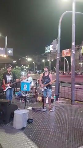 A low-angle, steady, full shot shows a band playing on a city sidewalk at night. The band consists of three men: a bassist on the left, a drummer in the middle, and a guitarist/singer on the right. The bassist is wearing an Oasis t-shirt and blue jeans. The drummer is wearing a light blue shirt. The singer is wearing a black tank top and jean shorts. A large speaker is on the ground to the left of the bassist, and a metal trash can is next to it. The drums are set up behind a blue box. A woman walks in front of the band from right to left at the beginning of the video. The sidewalk has a checkered pattern. In the background are city streets, traffic lights, and buildings. A sign indicates "Corrientes" and "1000". A Medife sign is visible on a building in the distance.