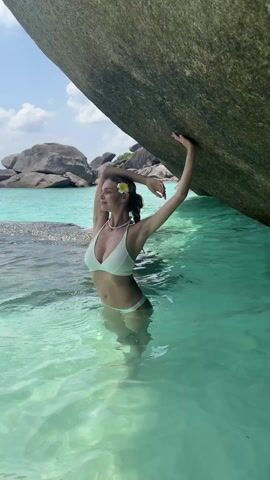The video shows a woman in a white bikini standing in clear water with a large rock to her side. The water is so clear, you can see the bottom of the ocean, and other rocks are scattered in the background. The woman poses with her arms raised, hands touching the rock. She has a white flower behind her ear and a necklace. She turns and smiles at the camera briefly before returning to her pose.