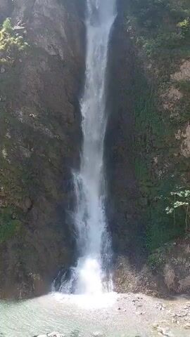 A tall waterfall cascades down a rocky cliff face covered in lush green moss and vegetation. The water plunges into a clear pool at the bottom, creating a misty spray. The sun shines brightly, illuminating the scene and casting shadows on the rocks. The surrounding environment appears to be a natural, undisturbed landscape.