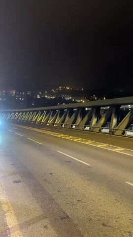 A red car approaches and passes a camera positioned on the side of a road at night. The road is wet and illuminated by streetlights and the car's headlights. The car's headlights are bright and cast reflections on the wet road. The car is moving from left to right in the frame and is blurred due to its speed. In the background, a bridge structure with illuminated supports and lights from a distant city are visible. The sky is dark and clear.