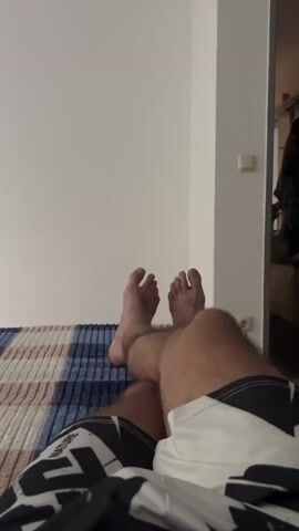 The video shows a first-person perspective of a person lying on a bed. Their legs are crossed, and their feet are visible, resting on a plaid blanket. The person is wearing patterned shorts. The background is a white wall and a corner of a room with a doorframe. The lighting appears to be natural or from a room light.
