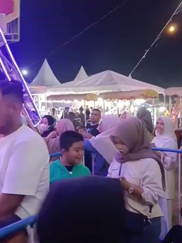 The video shows a crowded nighttime scene at what appears to be an amusement park or fair. In the foreground, several people are visible, including a man in a white t-shirt with a short haircut and a beard, looking towards the left. To his right, a young boy in a green and gray jacket is looking down at a phone. Further to the right, a woman in a light-colored hijab is also looking at a phone. Behind them, a Ferris wheel-like structure is illuminated with colorful lights, predominantly green, yellow, and purple. People can be seen boarding or disembarking from the ride's compartments, which are arranged in a circular pattern. Several tents or stalls are set up in the background, suggesting a fairground environment. The overall atmosphere appears lively and festive, with many people dressed in modest attire, indicating a likely cultural event. The lighting is dim, with the bright lights of the amusement ride and vendor stalls providing most of the illumination.