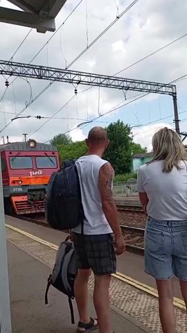 The video begins with a rear view of a man and a woman standing on a train platform. The man, bald and wearing a white sleeveless shirt and plaid shorts, has a large backpack slung over his shoulder. He also has a second backpack at his feet. The woman, with blonde hair and wearing a white t-shirt and denim shorts, stands next to him. A red train engine with the word "Евпатория" visible on its side is parked on the tracks to their left. The train has its headlights on. The camera then pans to the right, showing the side of the train as it begins to move. The train cars are gray and red. The camera continues to follow the moving train, showing its wheels and the tracks. The scene appears to be at a train station on a cloudy day.