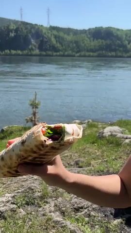 A person is seen holding and eating a large wrap or shawarma in a scenic outdoor setting. The wrap is golden-brown, likely grilled, and reveals fillings of vibrant green vegetables, red sauce or tomatoes, and other ingredients.

In the initial and final shots, a hand presents the wrap, showcasing its contents. In the middle shot, a person, viewed in profile, wearing a black hat and a black top, takes a large bite from the wrap.

The background features a wide, blue-green river flowing under a clear blue sky. On the opposite bank, lush green hills or mountains, densely covered with trees, rise up. Two tall power line towers are visible in the distance on the hills. The immediate foreground consists of rocky and grassy terrain. The scene depicts a sunny day, suggesting a pleasant outdoor meal experience in a natural environment. The location is Sliznevo, Russia.