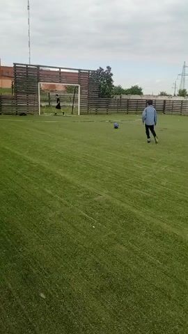On a green artificial turf field, a person in a blue shirt and dark pants kicks a soccer ball toward a goal, where another person stands, seemingly the goalie. The ball goes into the goal. The field is enclosed by a wooden fence. A building is visible in the background, as well as a cell tower. The sky is overcast.