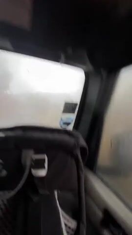 The video shows a view from the inside of a car, driving in very foggy and rainy weather. The windshield wipers are on, and the visibility is very poor, with a gray and blurred view of trees and landscape outside. The camera is inside the car, behind a car seat, focusing on the road ahead and the weather conditions. The interior of the car is visible, including the steering wheel, dashboard, and side window, all darkened due to the low light conditions and the focus on the outside view.