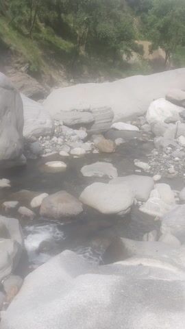 The video provides a top-down view of a shallow, rocky stream. The water is clear and appears to be flowing over smooth stones. The surrounding environment is rocky and undeveloped, with some vegetation visible on the left side of the frame. The lighting suggests it is daytime, and there is some fog or mist in the air, which gives the video a hazy appearance. The camera is moving slightly, creating a subtle sense of motion.