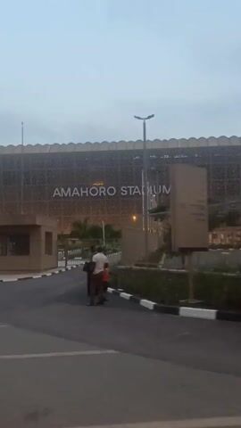 The video presents a medium shot of Amahoro Stadium in Rwanda, presumably in the evening, as suggested by the lighting. The stadium is a large building with an ornate, patterned exterior. The words "AMAHOOO STADIUM" are prominently displayed on the front of the stadium. In front of the stadium, there is a small, block-shaped guardhouse. A paved road leads to the stadium's entrance, and there are some trees and plants along the roadside. Initially, there are a few people walking near the guardhouse, but they move away. Later, a man walks across the road in front of the stadium. The sky is overcast, adding to the subdued atmosphere of the scene. Overall, the video provides a clear view of Amahoro Stadium and its immediate surroundings.