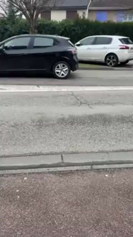 The video shows a residential street on an overcast day. A white Peugeot is parked on the side of the road, adjacent to a grassy area and a hedge. A brown Renault station wagon with a flat tire drives by. The car is travelling from right to left in the frame. The road has a crosswalk and is made of asphalt. The camera appears to be handheld and remains static throughout the video. The surrounding area includes houses with a variety of architectural styles and color schemes.