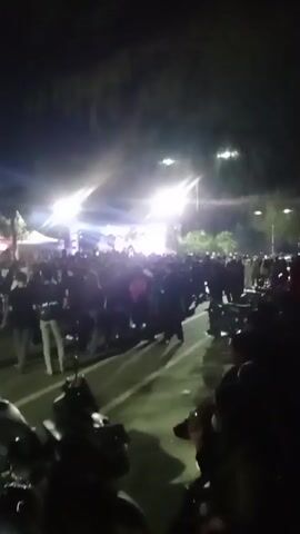 The video shows a large crowd of people gathered at night, possibly at an outdoor event. Bright stage lights are visible in the background, illuminating a stage area with tents set up behind it. The crowd is dense, with many individuals standing and observing the stage. Several motorcycles are parked in the foreground, suggesting attendees arrived by vehicle. The overall atmosphere appears to be that of a concert or a similar public gathering.