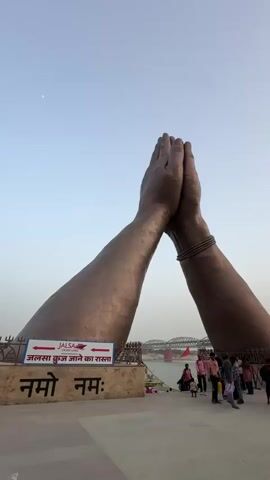 The video shows a large statue of hands in a prayer pose, with a bridge in the background and people walking around the statue. The hands are bronze in color, and there's a sign in front of the statue with text in what appears to be Hindi or Sanskrit. People of various ages and clothing styles walk in front of the statue. The sky is clear with a pale color gradient, suggesting dawn or dusk.