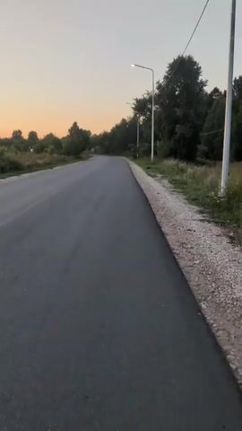 The video shows a paved road with a gravel shoulder, lined with trees and vegetation. A street lamp is visible on the right side of the road. The sky is a gradient of orange and yellow, suggesting it is either sunrise or sunset. The camera is moving, appearing to be handheld and tracking the road ahead. There are no people or vehicles visible in the frame.