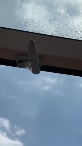 A video shot from below looking up at a partially opened skylight on a cloudy day. The skylight has a white handle and a wooden frame. Raindrops are visible on the glass. The sun is peeking through the clouds, casting a bright glare on the skylight.