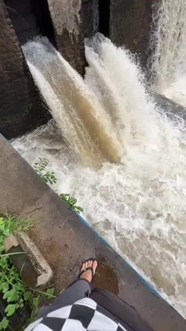 This media file depicts a powerful water release from a dam. The primary subjects are the rushing water and the concrete and stone structure of the dam. Two large pipes are actively discharging murky water downwards with significant force, creating a turbulent and frothy pool below. The dam itself is made of rough-hewn stones, with concrete elements visible. The surrounding environment includes lush green vegetation on the left bank and a dense treeline with palm trees in the background, suggesting a tropical or subtropical location. A distant bridge is also visible over the water.

The activity taking place is the controlled release of water from the dam, indicating a high water flow scenario. The time of day appears to be overcast, as there is no direct sunlight visible and the light is diffused. The weather seems to be humid, consistent with a tropical climate.

Notable details include the vibrant white foam and spray generated by the falling water, highlighting its energy. The water itself is a cloudy brown, suggesting it is carrying sediment. The perspective is from a slightly elevated position, looking down at the water flow. The image evokes a sense of raw natural power and the engineering prowess required to manage it. No visible text is present in the media file. The location context provided is Klungkung, Indonesia, which aligns with the visual cues of lush vegetation and the style of construction.