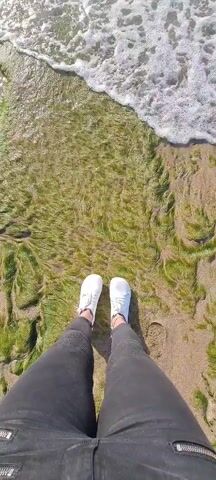 The video shows a person standing on a beach covered in green seaweed. They are wearing gray jeans and white sneakers. The ocean water washes over the seaweed near the person's feet. The camera angle is from a first-person perspective, looking down at the person's legs and the ground. The scene appears to be outdoors on a sunny day.
