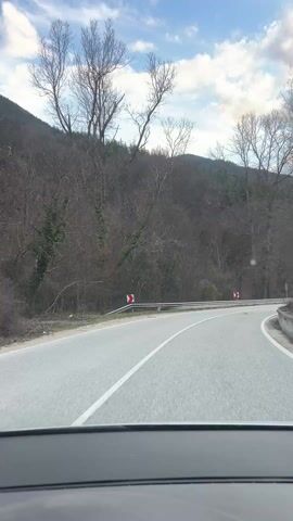The video captures a scenic drive along a winding asphalt road through a hilly or mountainous landscape. The perspective is from inside a vehicle, looking out through the windshield, with the top of the dashboard visible at the bottom of the frame.

The road features white lines, both solid and dashed, indicating lanes and the edge of the road. A metal guardrail runs along the right side of the road for much of the video. Several red and white chevron signs, indicating sharp curves, are visible along the guardrail. At the 0:03 mark, a triangular warning sign with an exclamation mark is also seen on the right side of the road.

The surrounding environment consists of hills densely covered with bare trees, suggesting it is either winter or early spring. Some of the trees have ivy growing on their trunks. The sky is partly cloudy, with patches of blue and dramatic formations of white and grey clouds. Sunlight occasionally breaks through the clouds, creating a dynamic lighting effect.

As the video progresses, the car navigates several curves, primarily to the left, as indicated by the road and the chevron signs. The overall impression is of a tranquil journey through nature. Background music is audible throughout the video.