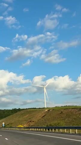 The video shows a highway with a wind turbine visible on a grassy hill in the background. The sky is blue with scattered white clouds. The road has multiple lanes and guardrails. Several vehicles, including cars and a truck, are driving on the highway. The wind turbine is white and has three blades, which are slowly rotating. The hill is covered in grass and some sparse vegetation. The overall scene depicts a scenic landscape with a focus on renewable energy infrastructure.