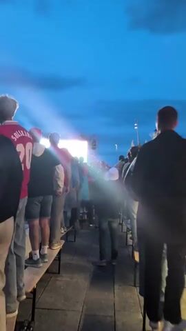 The video shows a crowd of people standing and sitting on benches, looking towards a large illuminated screen. The sky is dark with clouds, suggesting it is either dusk or night. There is a bright light emanating from the screen, which appears to be a large display or stage. Some people are wearing red shirts, and one person in the foreground has a red shirt with the number 20 on the back. The atmosphere seems to be one of a public gathering, possibly for an event like a concert, movie screening, or sporting event. The camera is moving, creating a slightly shaky perspective.