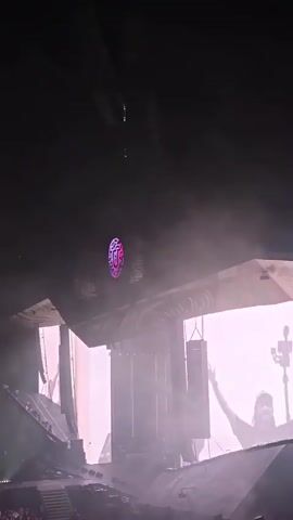 The video shows a large, elaborate stage setup with dynamic lighting and pyrotechnics during a live performance. The stage features multiple screens displaying colorful visuals, complex structures that resemble abstract shapes or architecture, and what appear to be speakers hanging from above. There are bursts of flame and smoke emanating from various points on the stage, creating a dramatic and energetic atmosphere. The lighting shifts between deep purples, pinks, and oranges, illuminating the haze and contributing to the visual spectacle. In the background, a silhouette of a person holding a staff or pole is visible, suggesting the presence of performers or stage crew. The overall impression is of a large-scale concert or festival event with impressive stage design and special effects.