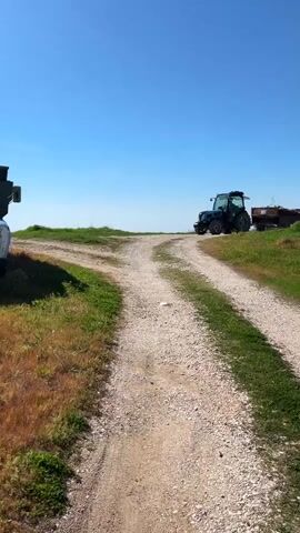 The video shows a rural scene with a gravel road that splits into two paths. On the right path, a blue tractor pulls a trailer loaded with debris or scrap. The tractor and trailer are moving away from the camera, disappearing over a slight rise in the landscape. To the left of the road split, part of another piece of farm machinery is visible. The sky is clear and blue, and the roadside is covered with green grass and some patches of dry vegetation. The overall scene suggests an agricultural or rural setting.