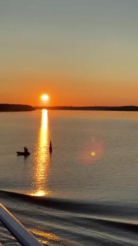 A video captures a sunset over a body of water. The sun, a bright orange orb, is low on the horizon, casting a shimmering golden reflection on the water's surface. The sky transitions from a pale yellow near the horizon to a soft gradient of orange and then to a muted blue-gray at the top. Silhouetted against the vibrant sky is a tree-lined landmass on the left and a distant, darker landmass on the right, possibly with a faint tower. In the water, a small, dark boat with two figures is visible to the left of the sun's reflection. A red buoy also stands in the water to the right of the boat. The foreground shows a portion of a white railing, suggesting the viewer is on a vessel. The water exhibits gentle ripples, particularly in the foreground, indicating movement. The overall scene is tranquil and depicts the end of the day.