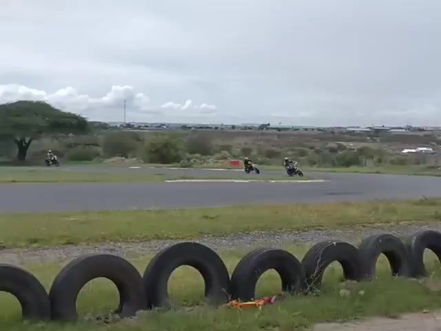 The video shows three motorcycles racing on a paved track. Each rider wears a helmet and racing gear. The track is surrounded by grass and a row of black tires at the forefront. In the background is a landscape of shrubs, sparse trees, and a distant cityscape under a cloudy sky. The motorcycles round a curve in the track during the race.
