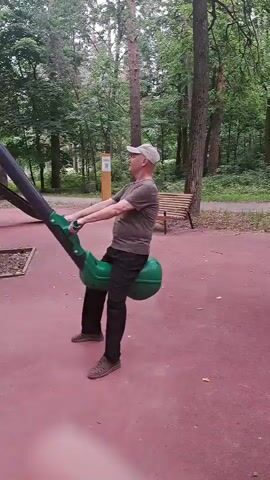 An elderly Caucasian man wearing a grey t-shirt, black pants, a beige baseball cap, and beige shoes is sitting on a green seesaw in a park. He is holding onto the handles of the seesaw, which is in motion. The man is facing the right side of the frame, and his head is tilted slightly upwards. The seesaw is located on a red rubberized surface, and the background consists of trees and a wooden bench. The lighting suggests it is daytime, and the environment appears to be a recreational area.