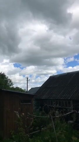 A vertical video shows a rustic scene with a wooden outhouse in the foreground. The outhouse is dark brown and appears weathered. To the right of the outhouse is a larger, dark-roofed building, possibly a house or barn, with visible windows. Behind these structures, a line of trees and bushes forms the middle ground. A tall evergreen tree stands to the far left. The background is dominated by a dramatic sky filled with heavy, gray clouds interspersed with patches of blue. A power pole is visible in the distance between the trees and the larger building. The overall impression is of a rural setting on a cloudy day.