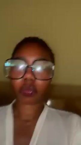 A person wearing oversized glasses and a white shirt smiles at the camera. The background is a beige wall and a bed headboard. The person briefly looks to the side before the video ends.