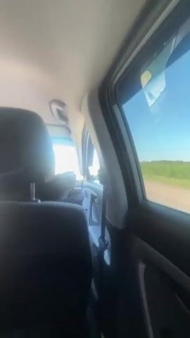 A person is inside a moving vehicle, filming out of the passenger window. The scene outside shows a road with grass and trees on either side. The sky is bright blue.  A utility pole can be seen in the distance.  The interior of the car features a black seat and trim.  The window is slightly tinted.