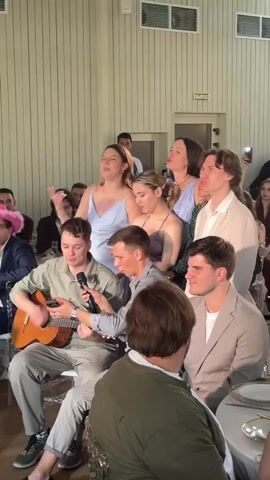 A group of young adults are gathered indoors, possibly at a formal event or party. In the foreground, a young man with short brown hair, wearing a light green collared shirt and grey pants, is seated and playing an acoustic guitar. He is also holding a microphone close to his mouth, suggesting he is singing. Beside him, another young man with short brown hair, wearing a grey shirt, is looking towards the guitarist. To the right of the guitarist, a young man with brown hair, wearing a light brown suit jacket, is looking forward. Behind these seated men, a group of four young women are standing, singing with their mouths open. They are wearing dresses; one in light blue, one in purple, and one in a blue and green patterned dress. A young man in a light cream suit jacket stands behind them. In the background, other guests are seated at tables, and a man in a suit is visible near a doorway. The environment appears to be a room with pale green paneled walls and a white ceiling with lights.