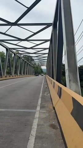 This media file is a first-person perspective video clip showing a view from a bridge. The bridge is a steel truss bridge with a concrete road surface. On the right side of the road, there is a yellow concrete barrier with black chevron markings. The barrier is segmented, indicating it's likely a traffic barrier.

The road itself has white lane markings and textured concrete for pedestrians or cyclists. Several vehicles are visible on the bridge: a black SUV, a motorcycle, and another car further in the distance. The scene suggests traffic is moving across the bridge.

The surrounding environment includes lush green trees and vegetation on either side of the bridge, with a body of water, possibly a river, visible below the bridge on the right. In the background, hills or mountains can be seen, partially obscured by trees and clouds.

The sky is overcast, indicating a cloudy or perhaps a slightly hazy day. The time of day appears to be daytime, given the ambient light. There is no visible text in the media file. The overall mood is neutral, conveying a sense of travel or transit. The location context provided is Lamkruet, Indonesia.