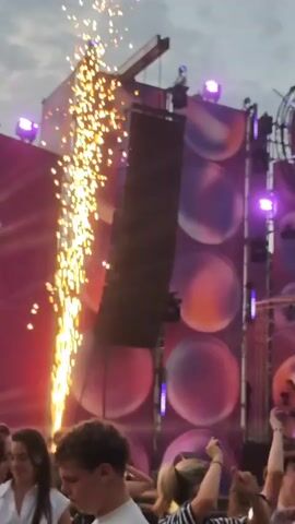 The video shows a crowded outdoor music festival. A large stage is set up with a prominent circular sign that reads "purple" in white lettering against a purple background. The stage is adorned with large banners featuring abstract purple and orange circular designs. Several people are visible on stage, some holding microphones and appearing to perform. Bright purple lights illuminate the stage and the surrounding area. A large speaker system is positioned to the left of the stage. In the foreground, a crowd of people are gathered, many facing the stage and some holding up cell phones to record the event. The overall atmosphere suggests a lively and energetic musical performance. The sky is cloudy.
