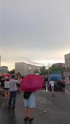 A group of people stand on a wet street, some holding umbrellas, as a water truck sprays a stream of water into the air. The sky is overcast and the lighting suggests it is late afternoon or early evening. In the background, apartment buildings are visible. Some people are holding up phones, possibly recording the event.
