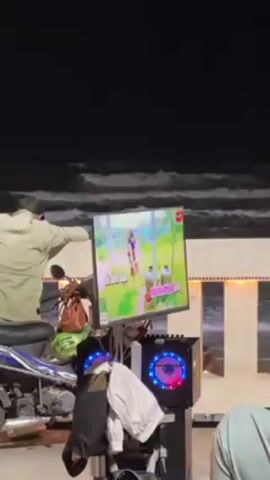 A group of people are enjoying karaoke at an outdoor venue. A man stands to the side near a parked motorbike, while another man faces away from the camera, possibly watching the performance. The karaoke system includes a monitor displaying lyrics and visuals, and a speaker with a light display. In the background, a structure designed to mimic a beach scene includes a painted backdrop of waves. People stand on a raised platform singing into a microphone. The scene is at night and illuminated by lights.