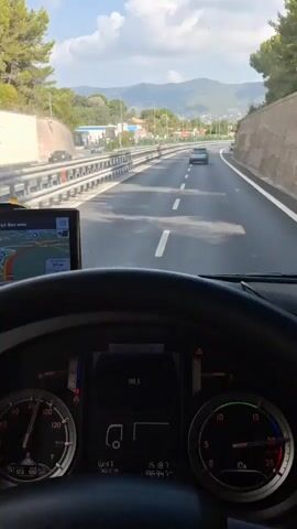 The video shows the interior of a truck's cabin. The driver's perspective is looking forward through the windshield. A steering wheel is visible in the foreground, with a dashboard displaying various gauges and a digital screen. A GPS device is mounted to the left of the steering wheel, showing a navigation route. The truck is on a highway, with a concrete barrier to the left and a sloping embankment to the right. Other vehicles can be seen on the road ahead and to the left. The landscape outside includes trees, buildings, and mountains in the distance under a cloudy sky.