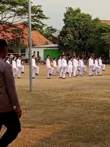 A group of young people in white uniforms are performing a synchronized march on a grassy field. They are spaced in rows and appear to be practicing some form of drill or dance. A person in a police uniform is standing in the foreground, facing away from the camera and towards the group. The environment is outdoors with trees and buildings in the background. The sky appears to be overcast. The camera is positioned at a slightly elevated angle.