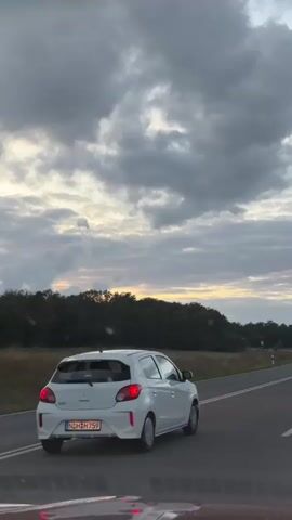 A white car drives on a paved road through a rural landscape under a cloudy sky. The car is in the foreground, followed by another vehicle from which the video is filmed. The road curves to the right, with trees and fields on either side. The sky is filled with dark gray clouds, with a hint of yellow and orange from the setting sun peeking through.