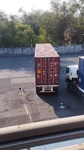 A long, dark red shipping container is being transported on the back of a white truck. The container has various labels and markings on its side. Another truck, white with a blue container, is parked to the right of the first truck. The scene takes place outdoors in what appears to be a port or industrial area, with a fence and trees in the background. The ground is paved asphalt. The sun is shining.