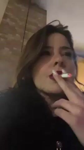 A woman with brown hair, red lipstick, and white fingernails is smoking a cigarette outdoors. She takes a drag from the cigarette and exhales the smoke. The background consists of a brick wall and dimly lit surroundings. She wears a black jacket.