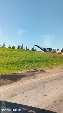 The video shows a view from inside a vehicle traveling on a dirt road. The road is on the left side of the frame, and the right side of the frame shows a grassy hill with scattered trees and some construction equipment. The sky is clear and blue. There are also some power lines visible on the hill. The vehicle's window reflects the sky and the scenery. The video continues to show the view from the car as it drives. The construction equipment is still visible in the first few seconds. Later, the scene changes to a dirt lot with more construction vehicles. Then it goes back to the grassy hill with power lines. The sun is bright and casts shadows. The video is shot from a moving vehicle.